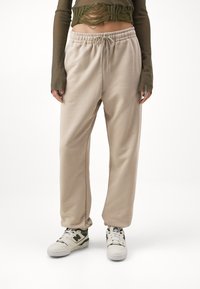 Beige Jogginghosen aus weichem Stoff mit elastischem Bund und Kordelzug. Getragen mit einem grünen Oberteil und weißen Schuhen mit grünen Akzenten.