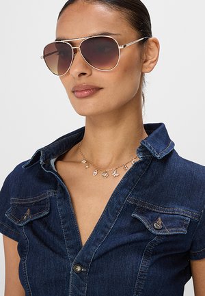 Donna che indossa grandi occhiali da sole aviator, una camicia di jeans blu con colletto aperto e una delicata collana d'oro con ciondolo.
