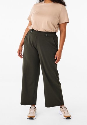 Mujer con camiseta beige de manga corta, pantalones anchos de pierna en verde oscuro con detalles de botones y zapatillas beige sobre fondo blanco.