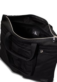 Svart stoff tote bag med solide håndtak, glidelås-lukking, kontrasterende hvite sømmer, og en innvendig etikett med merkeinitialene "CK."