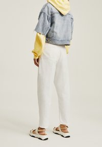 Chaqueta de mezclilla corta, sudadera con capucha amarilla, pantalones blancos de corte holgado, sandalias beige con suela negra, mostrando un look casual en capas.