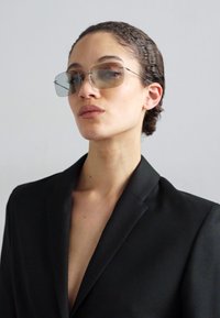 Blazer noir avec un décolleté en V profond, associé à des lunettes de soleil légèrement teintées. Texture de tissu lisse et design structuré. Esthétique minimaliste.
