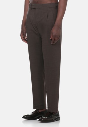 Uomo che indossa pantaloni marrone scuro su misura con risvolti e mocassini in pelle nera, in piedi davanti a uno sfondo chiaro e uniforme.