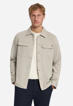 Mann mit kurzem welligem blondem Haar, trägt eine hellbeige Knopfleistenjacke über einem weißen Hemd und dunkler Hose, Hände in den Taschen, neutraler Hintergrund.