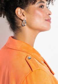 Veste en daim orange avec épaulettes et quincaillerie en argent. Le modèle porte de grandes boucles d'oreilles en argent brillant. Texture lisse, angle rapproché.