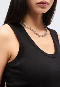 Νεαρή γυναίκα που φοράει μαύρο αμάνικο ριμπ μπλουζάκι και ασημένιο κολιέ με αλυσίδα σε σχήμα αστεριού και κόμπους.