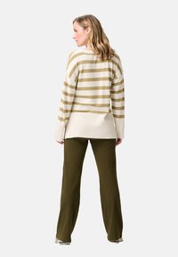 Vrouw die wegkijkt en een beige en wit gestreept trui draagt met olijfgroene ribbelbroek en lichte sneakers.
