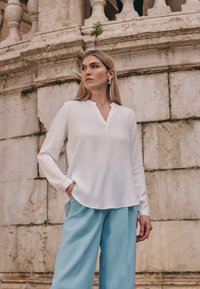Weiße Langarmbluse mit V-Ausschnitt und abgerundetem Saum, kombiniert mit hellblauen weitem Hosen. Glatter Stoff, minimalistisches Design.