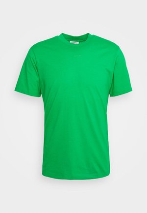 Man draagt een felgroen shirt, een zwart, kort mouwen, knoopjasje, witte broek met trekkoord en zwarte leren schoenen tegen een witte achtergrond.