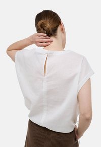 Haut en lin blanc avec une ouverture en forme de clé dans le dos, des manches courtes et une coupe décontractée. Le tissu présente une texture subtile.