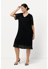 Mujer vestida con un vestido de encaje negro hasta la rodilla con mangas cortas y sandalias negras de tacón alto con lazos joya, sonriendo y tocándose la frente.
