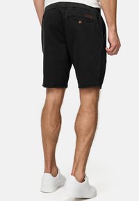 Pantalones cortos de algodón negros con cintura elástica, bolsillo trasero y un parche de cuero. Combinados con zapatillas blancas, mostrando un corte a la altura de la rodilla.