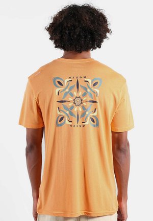 Homme aux cheveux bouclés portant un t-shirt jaune moutarde avec un motif floral symétrique bleu et beige et le texte « Oxbow » au dos.