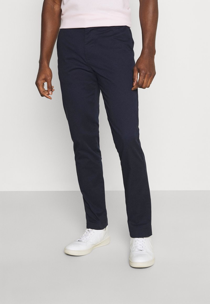 Homme portant un pantalon slim bleu marine foncé et des baskets blanches, debout devant un fond clair uni.