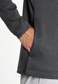 Vêtement gris à manches longues avec un tissu texturé, des poches zippées sur les côtés et une coupe décontractée. Les poignets sont terminés par des bords cousus propres.