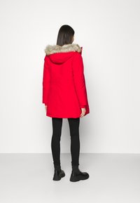Parka roja con capucha de piel sintética, que cuenta con cierre de cremallera y botones a presión. Tejido texturizado y un diseño de largo hasta el muslo. Combinada con leggings negros y botas.