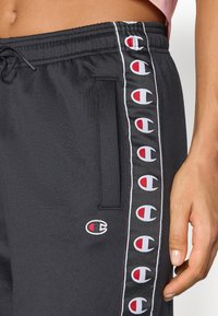Calças de jogging pretas feitas de um tecido macio, com bolsos laterais, cintura elástica e riscas laterais com logotipo repetido em vermelho e branco.