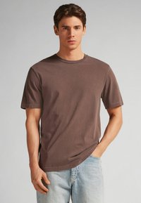 Hnědé bavlněné tričko s krátkým rukávem, kulatým výstřihem a uvolněným střihem, doplněné o světle modré džíny.