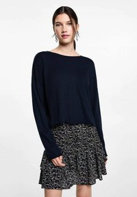 Marineblauer Strickpullover mit weitem Ausschnitt, kombiniert mit einem gemusterten schwarzen Rock, der florale Akzente und einen rüschenbesetzten Saum aufweist.