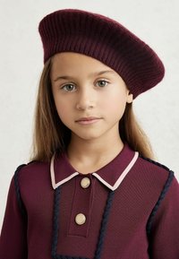 Berretto a maglia bordeaux con texture a coste, indossato di lato. L'outfit presenta un top bordeaux con un colletto rosa e bottoni dorati.