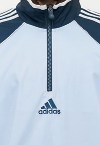 Λευκό αθλητικό μπουφάν με ναυτικές λεπτομέρειες, ψηλό γιακά, φερμουάρ μέχρι το ύψος του στήθους και σχέδιο με τρεις ρίγες στους ώμους. Διαθέτει κεντημένο το λογότυπο της Adidas.