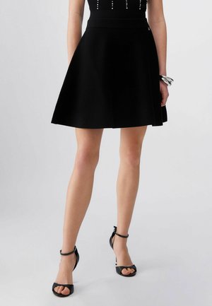 Femme portant une jupe noire évasée, des sandales à talons hauts avec bride à la cheville noires, et des bracelets argentés au poignet droit, sur un fond uni.