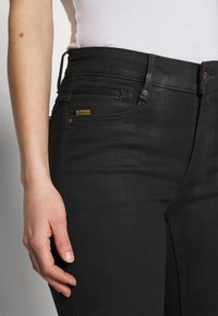 Vaqueros de mezclilla negra con cintura alta, diseño de cinco bolsillos y una etiqueta con el logo de G-Star. Textura suave con un sutil detalle de costuras verticales.