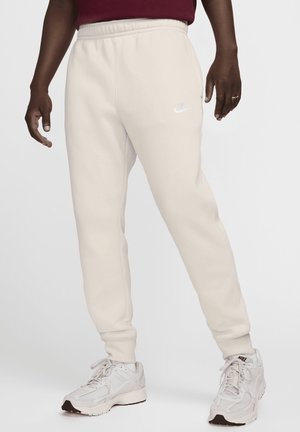 Unterkörper einer Person, die hellbeige Nike-Sweatpants und weiße Nike-Sneaker trägt, mit einem oberhalb der Taille sichtbaren dunkelroten Oberteil.