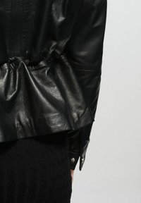 Czarna skórzana kurtka z marszczonym wcięciem w talii, posiadająca gładką fakturę i zapięcie na napę na mankietach. Czarna tkanina o fakturze poniżej.