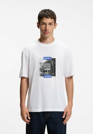 Ung mand iført hvid T-shirt med sort-hvidt naturfoto og tekst "Danger High BPM Zone Ahead" indrammet af blå "Hugo" og "Rave" mærkater.