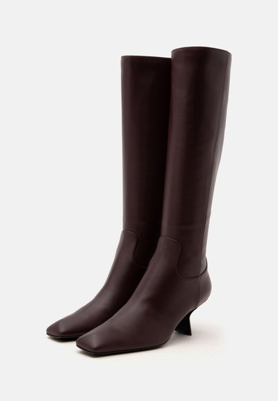 Paire de bottes en cuir brun foncé montantes jusqu'au genou, avec bouts carrés et talons bas courbés sur fond blanc.
