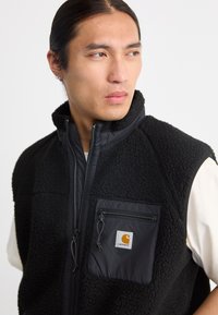 Melna vilnas veste ar rāvējslēdzēja priekšējo kabatu, kontrastējošu gludu auduma paneli un Carhartt logotipu uz kabatas. Augsta apkakles dizains.
