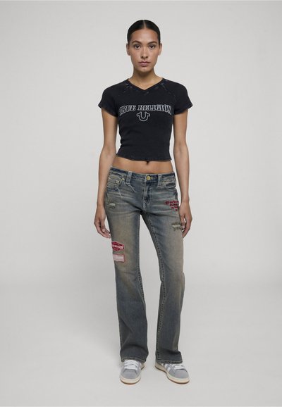Femme debout portant un crop top noir avec le texte « True Religion », un jean déchiré avec des patchs rouges et des baskets gris clair sur un fond uni.