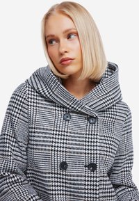 Zwart-wit houndstooth-patroon jas met een grote capuchon, dubbele knopen en een zachte, textuurstof.