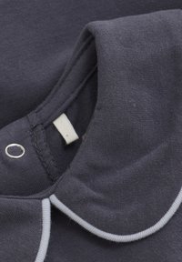 Marinblåt stof med blød tekstur, med en rund krave og lys grå piping. Inkluderer et mærke og en sølvfarvet trykknapdetalje.