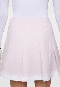 Kvinna som bär en ljusrosa plisserad kjol och en vit långärmad topp, visad från midjan till mitten av låret mot en neutral bakgrund.