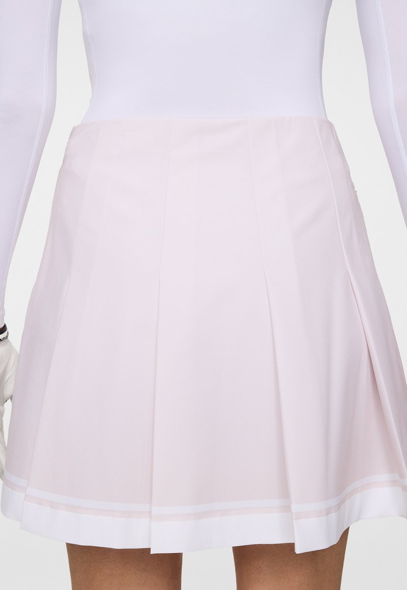 Frau in einem hellrosanen plissierten Rock und einem weißen Langarmoberteil, von der Taille bis zur Oberschenkelmitte vor einem einfarbigen Hintergrund abgebildet.