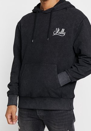 Mand iført en sort hættetrøje med teksten "Skully" på brystet og en forlomme, kombineret med mørke, revnede jeans.