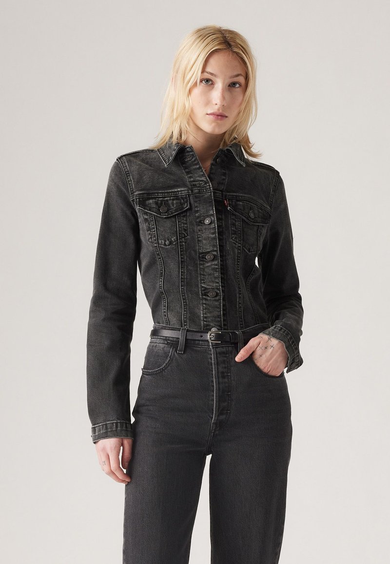 Junge Frau mit blonden Haaren, die eine enganliegende schwarze Jeansjacke und hochtaillierte schwarze Jeans mit einem schwarzen Gürtel trägt und vor einem schlichten Hintergrund steht.
