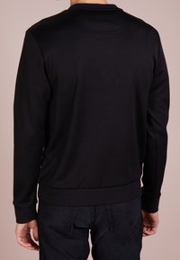 Uomo che indossa un semplice maglione a maniche lunghe nero e jeans scuri, visto da dietro su uno sfondo neutro.