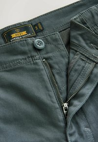 Közeli kép sötétzöld nadrágról, amelyen nyitva van a fémezett cipzár, a gomb bezárva, és a derékpánthoz csatolt "Next Durable Goods" márkás címke látható.