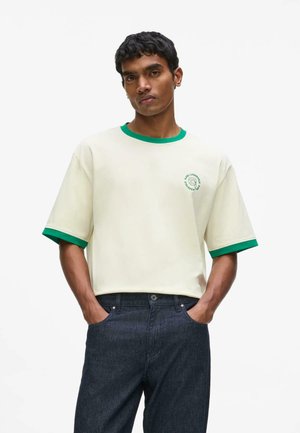 Homme portant une chemise écrue à manches courtes avec bordure verte et logo sur la poitrine, associée à un jean bleu foncé, mains dans les poches.