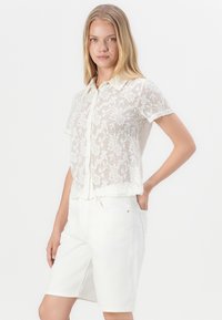 Blusa blanca de manga corta con un diseño de encaje floral y cuello clásico. Combinada con pantalones cortos de mezclilla de color claro.