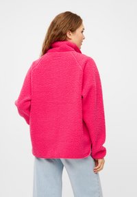 Giacca pullover in pile di colore rosa brillante con colletto alto, maniche a raglan e tessuto testurizzato. L'orlo presenta spacco laterale per facilitare i movimenti.