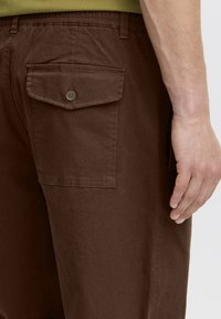 Pantaloni di cotone marrone con tasca posteriore chiusa da bottone e vita elastica. Vista ravvicinata che mostra la trama e i dettagli delle cuciture.