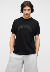 Νέος άνδρας με μεσαίου μήκους σκούρα μαλλιά, φορώντας μαύρο γραφικό μπλουζάκι και ανοιχτό γκρι σορτς, τα χέρια στις τσέπες, ουδέτερο φόντο.