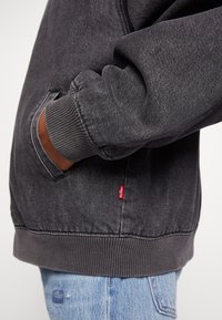 Personne portant un sweat-shirt Levi's gris foncé, main dans la poche, et un jean bleu clair déchiré visible à la taille.