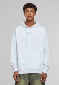 Sudadera blanca con cordones y un bolsillo tipo canguro, con texto bordado en verde en el pecho, llevada sobre unos jeans verde claro.