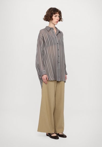 Camicia leggera e leggera grigia con strisce verticali bianche, con chiusura a bottoni e colletto, abbinata a pantaloni beige larghi e scarpe scivolate marroni scure.