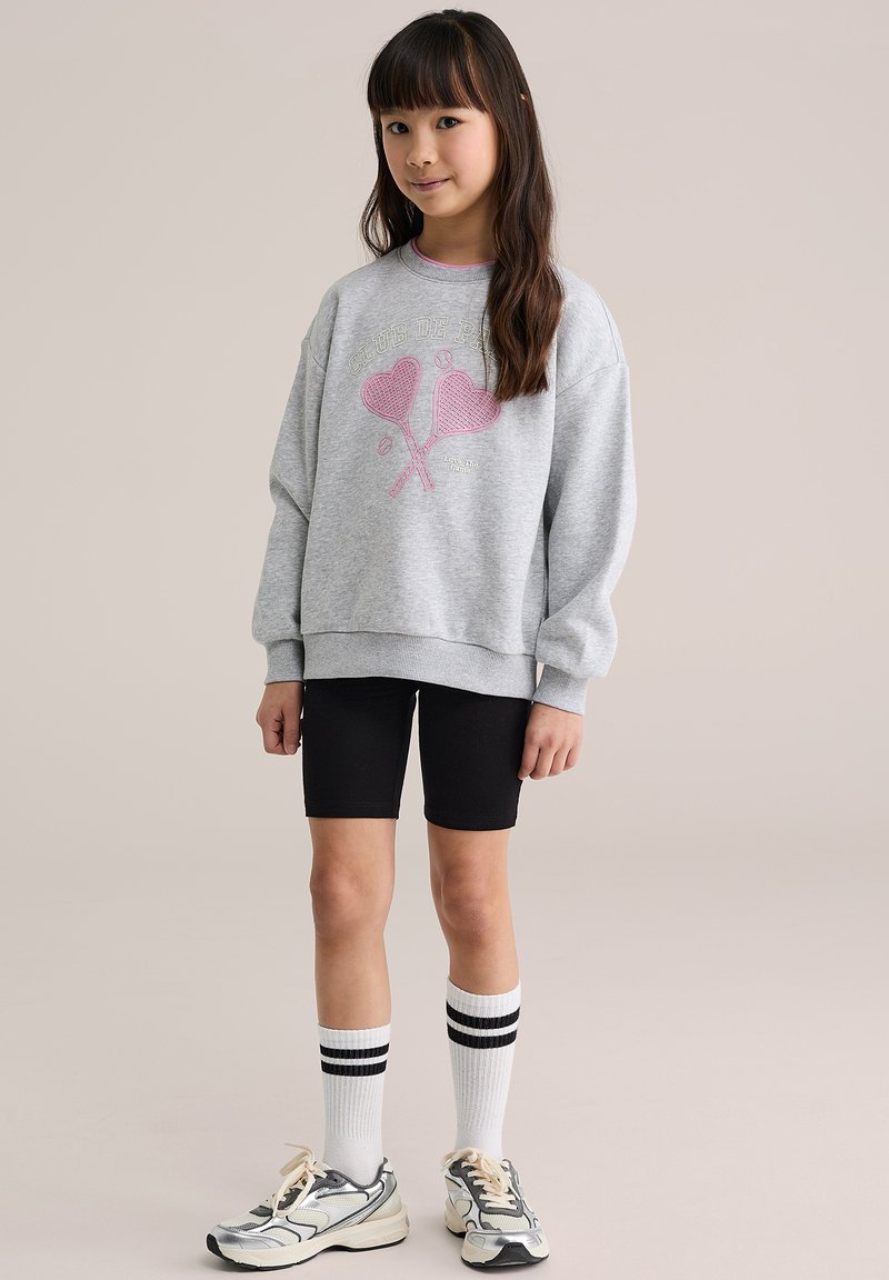 Halli pusa roosa südamepildiga, mustad rattasõidupüksid, valged põlveni sokid mustade triipudega ning hallid spordijalatsid beežide detailidega.